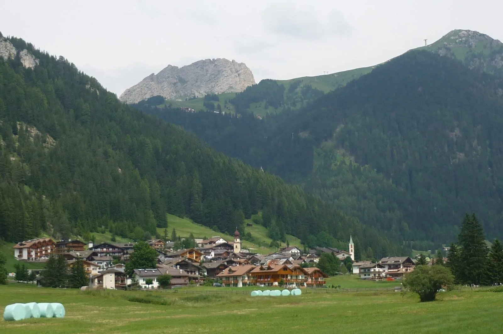 Veduta estiva di Canazei dai prati del fondovalle, con balle di fieno fasciate, case con balconi in legno, due campanili e montagne boscose sormontate da una cima rocciosa