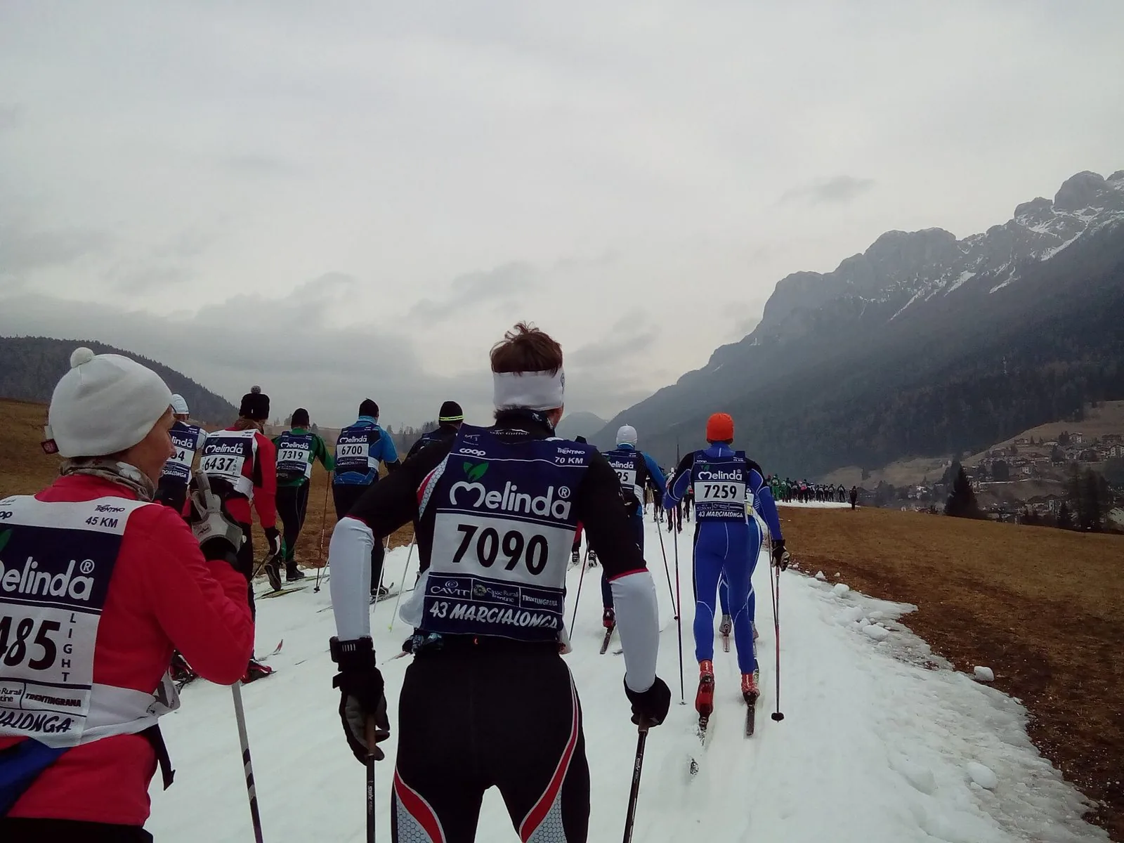 Fondisti con pettorali numerati della Marcialonga, ripresi di spalle, avanzano in fila su una pista di neve tra prati invernali, con montagne rocciose sullo sfondo