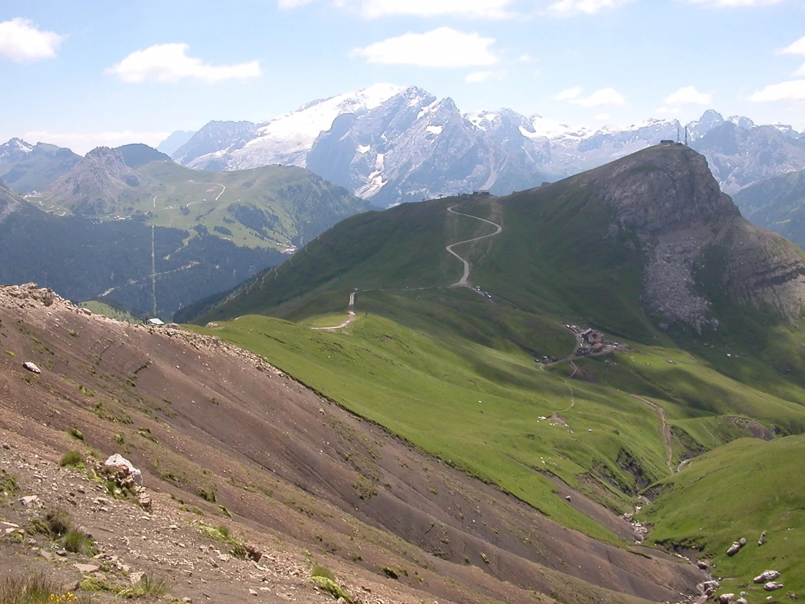 Montagne dolomitiche in estate: pendii erbosi con una strada sterrata a tornanti che sale verso il Col Rodella, a destra, e sullo sfondo il massiccio innevato della Marmolada sotto un cielo azzurro