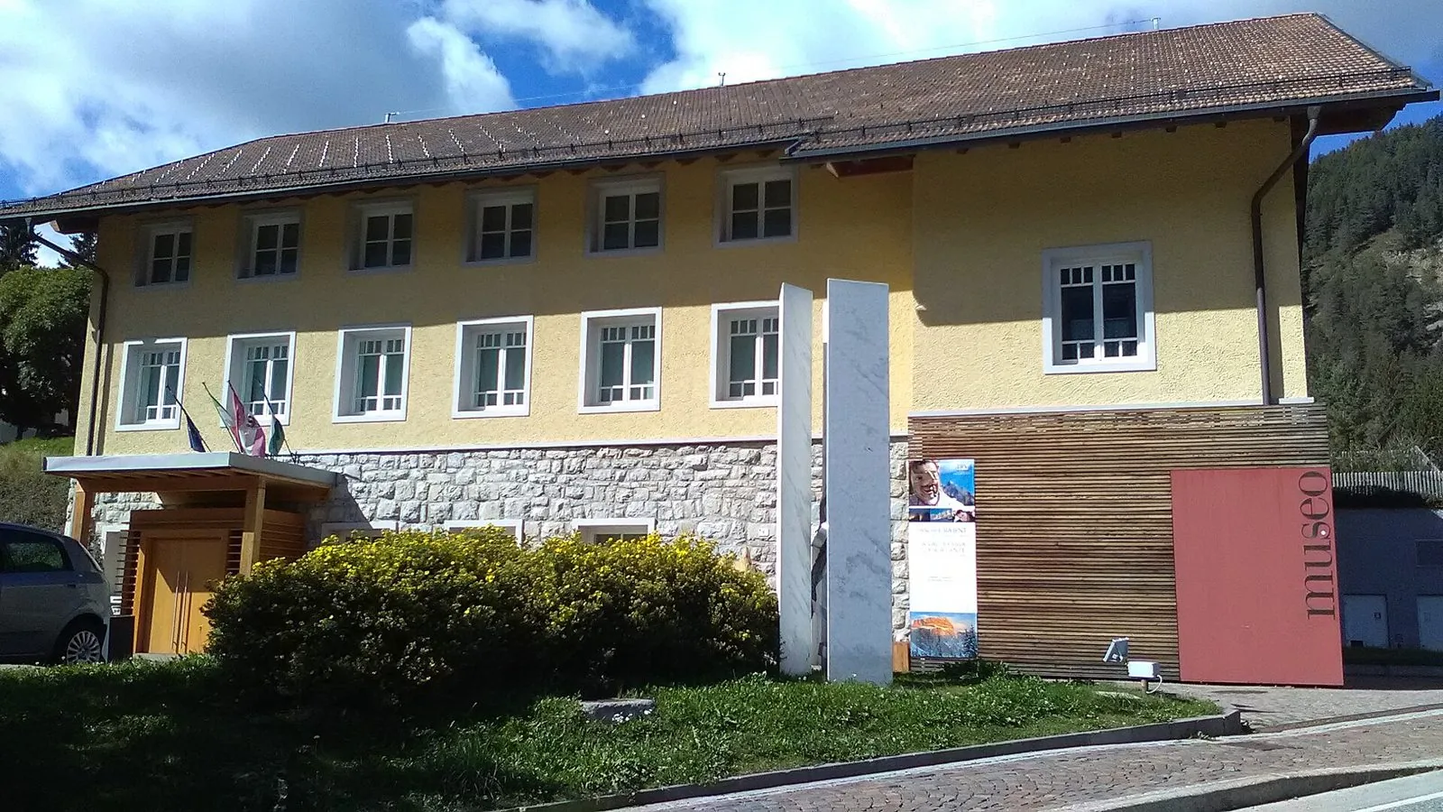 Facciata gialla a due piani del Museo Ladin de Fascia con basamento in pietra, finestre bianche, una parete rivestita in legno e un pannello rosso con la scritta museo, davanti a un'aiuola fiorita