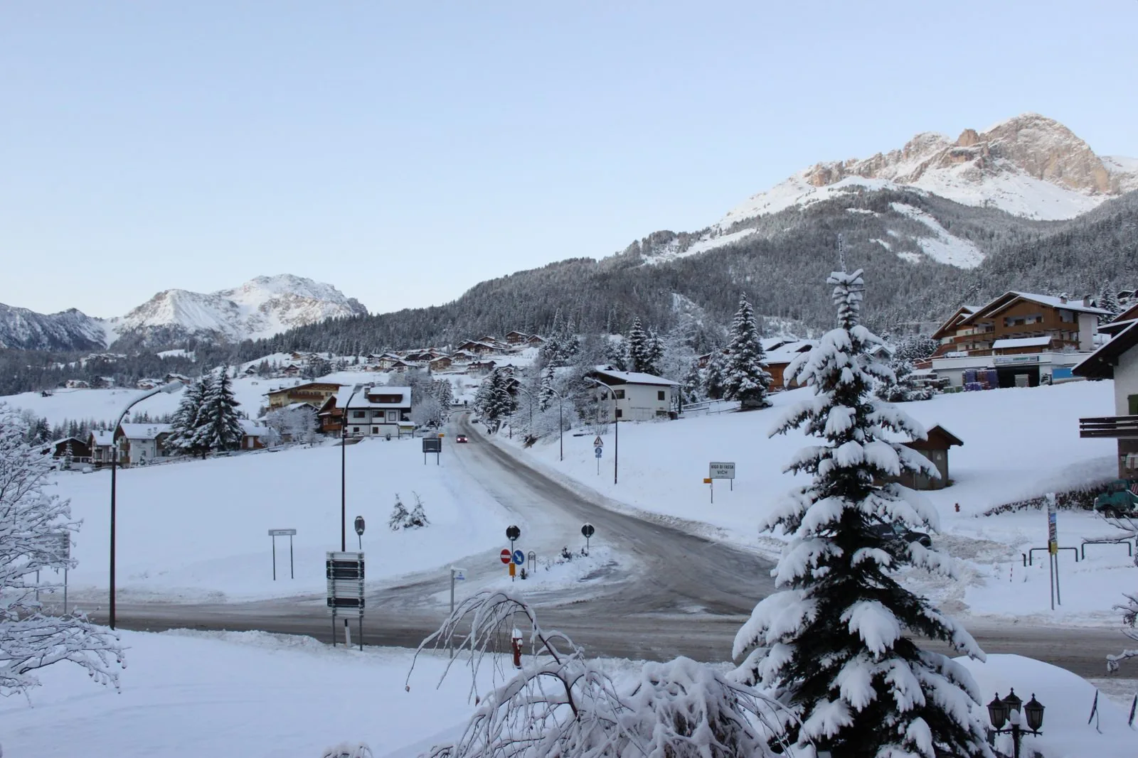 Strada che attraversa Vigo di Fassa dopo una nevicata, con abeti carichi di neve, case in stile alpino, cartelli stradali e cime innevate sullo sfondo